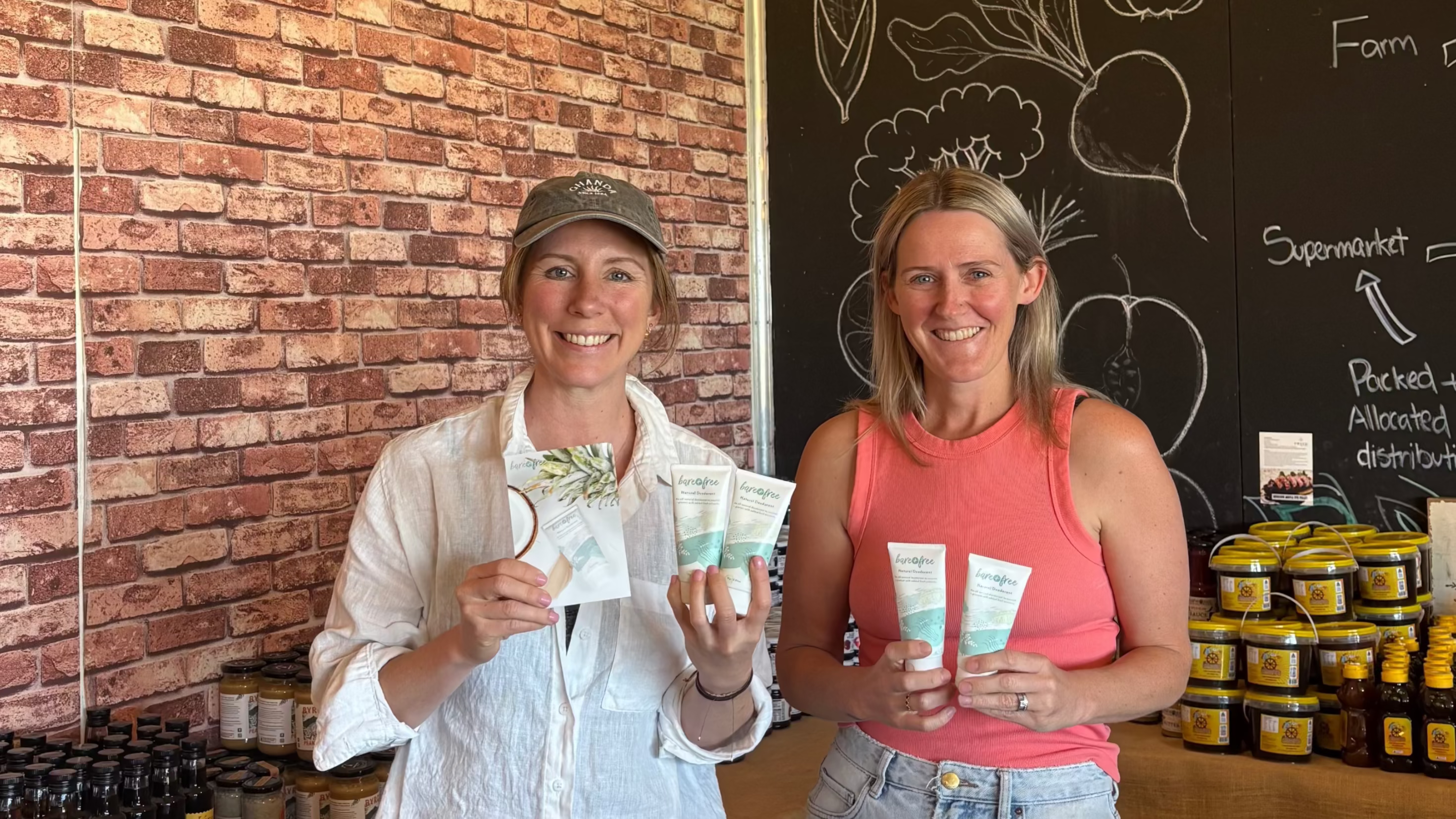 Two women standing in a store holding products, with a brick wall and chalkboard menu in the background.